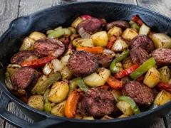 A cast-iron skillet of seared sausage coins, browned potato cubes, onion and strips of red and green pepper, on a weathered wooden table.