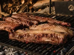 Thick cross-cut short ribs and a fat-capped flank on an Argentine parrilla grate, scattered with coarse salt over a bed of glowing embers.