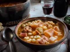 A bowl of thick Argentine locro — white corn, beans and collapsed squash — under a spoonful of red paprika fat and sliced scallion.