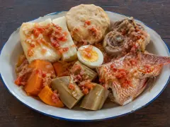 An enamel plate of boiled cassava, sweet potato and green banana with a halved boiled egg, a flat boil cake, a coil of pigtail and a piece of fried red snapper, all spooned with tomato and onion sauce.