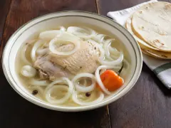 A bowl of clear broth with a piece of chicken, thick white onion rings, allspice berries and a whole orange habanero, with corn tortillas on a cloth beside it.