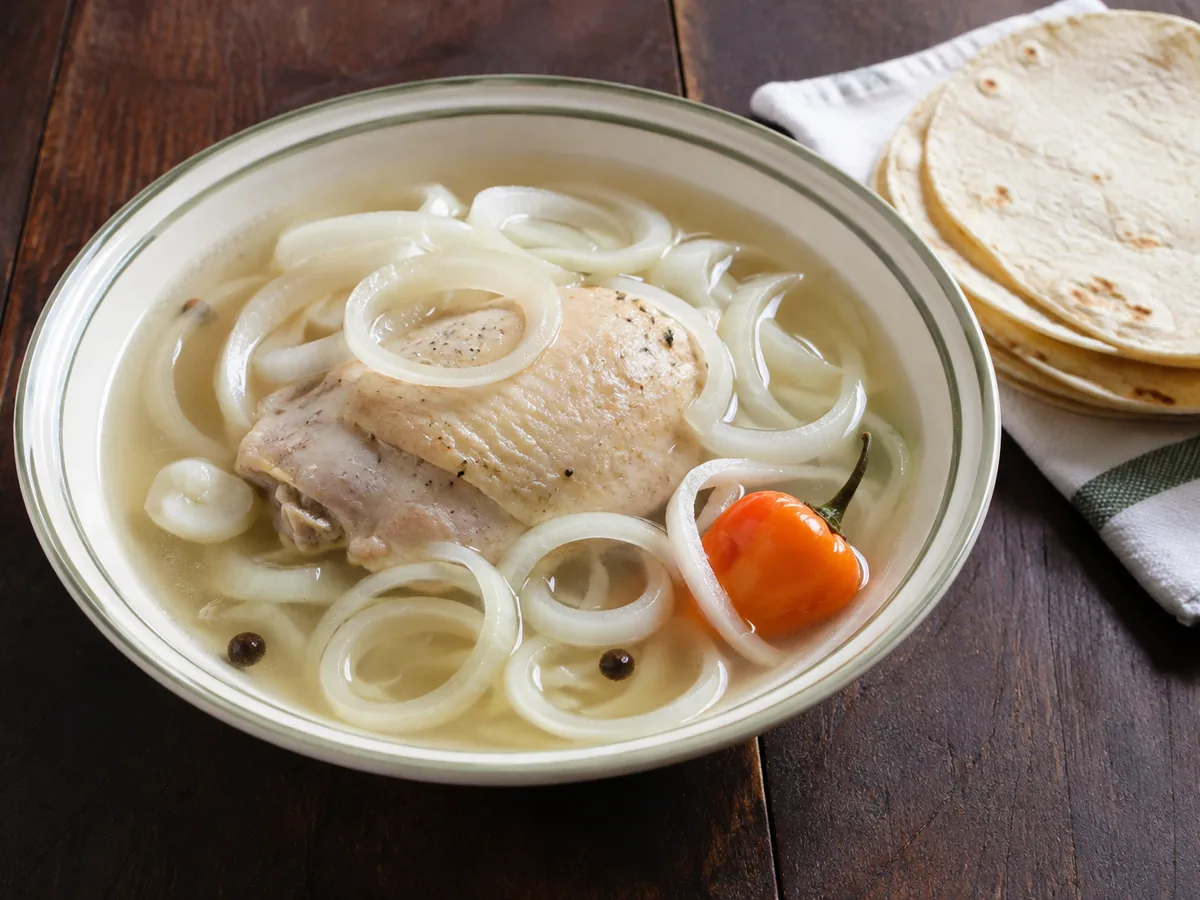 A bowl of clear broth with a piece of chicken, thick white onion rings, allspice berries and a whole orange habanero, with corn tortillas on a cloth beside it.