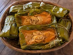 A basket of banana-leaf tamales tied with string, two of them opened to show orange-red masa with shredded chicken at the centre.