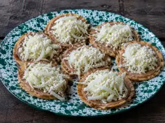 Seven crisp fried tortillas on a green-speckled enamel plate, each spread with refried beans and piled with grated white cheese and shredded cabbage and onion.