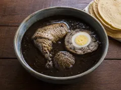 A blue-grey bowl of black broth holding a chicken leg, a slice of pork stuffing with a hard-boiled egg at its centre and a meatball, with a stack of corn tortillas behind.