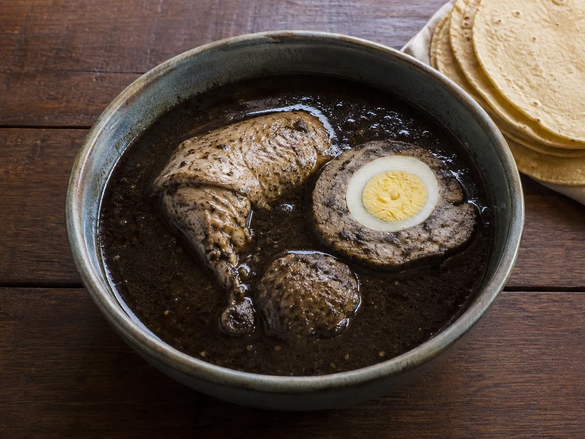 A blue-grey bowl of black broth holding a chicken leg, a slice of pork stuffing with a hard-boiled egg at its centre and a meatball, with a stack of corn tortillas behind.