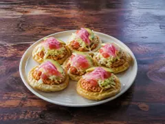 Six small puffed golden masa rounds on a plate, each topped with shredded orange chicken, cabbage, tomato, pink pickled onion and a slice of avocado.