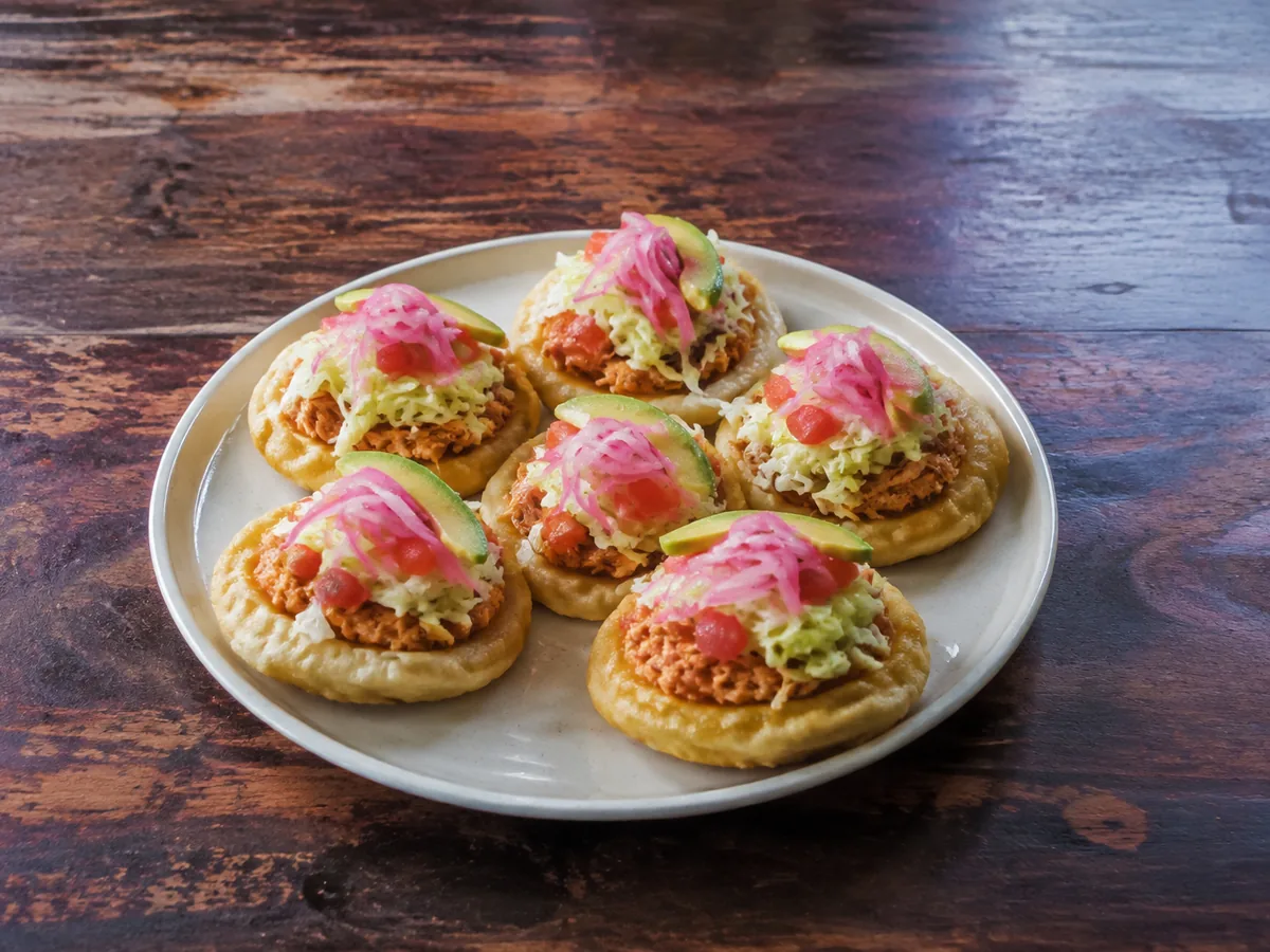 Six small puffed golden masa rounds on a plate, each topped with shredded orange chicken, cabbage, tomato, pink pickled onion and a slice of avocado.
