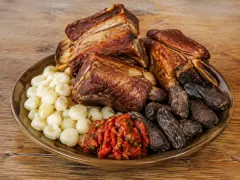 A wide olive-brown platter holding chestnut-brown fried pork rib chunks in the centre, a mound of large white hominy kernels on one side, dark wrinkled black chuño potatoes on the other, and a spoonful of red chunky chilli salsa at the front, on a wooden table.