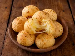 A shallow wooden bowl piled with round golden-crusted cheese rolls, the one at the front torn open to show a pale interior and a long strand of melted cheese stretching across the break, on dark wooden boards.