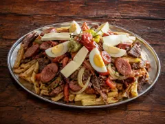 A large oval metal platter heaped with thick-cut chips buried under seared beef strips, sliced sausage rounds, onion and red pepper, topped with quartered hard-boiled eggs, white cheese batons and whole chillies, on a dark weathered wooden table.