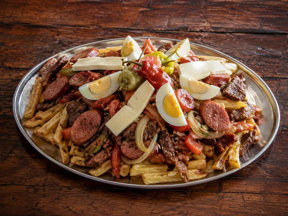 A large oval metal platter heaped with thick-cut chips buried under seared beef strips, sliced sausage rounds, onion and red pepper, topped with quartered hard-boiled eggs, white cheese batons and whole chillies, on a dark weathered wooden table.