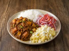 A pale ceramic plate quartered with a brick-red beef and potato stew studded with green peas, a mound of white rice, a heap of sliced tomato and red onion salad, and pale scrambled chuño flecked with crumbled white cheese, on a wooden table.