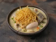 A dark clay bowl of pale cream-coloured soup holding a chicken piece, a beef rib, green peas and diced carrot, topped with a tall tangled nest of thin golden fried potato matchsticks, on a mottled dark surface.