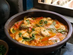 An orange-gold fish stew in a dark clay pot, with pepper strips, tomato rounds, and chopped cilantro over the top.
