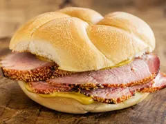 A tall sandwich of overlapping pink pork loin slices with golden cornmeal crusts on a soft Kaiser roll, mustard dripping down the side.