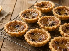 A dozen small butter tarts on a wire rack, their flaky pastry rims golden and their brown sugar fillings puffed and crackled, one broken to show a runny amber centre.