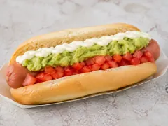 A long soft bun on a white paper tray holding a pink sausage under a band of finely diced red tomato, a band of mashed green avocado and a piped white crest of mayonnaise along its length.