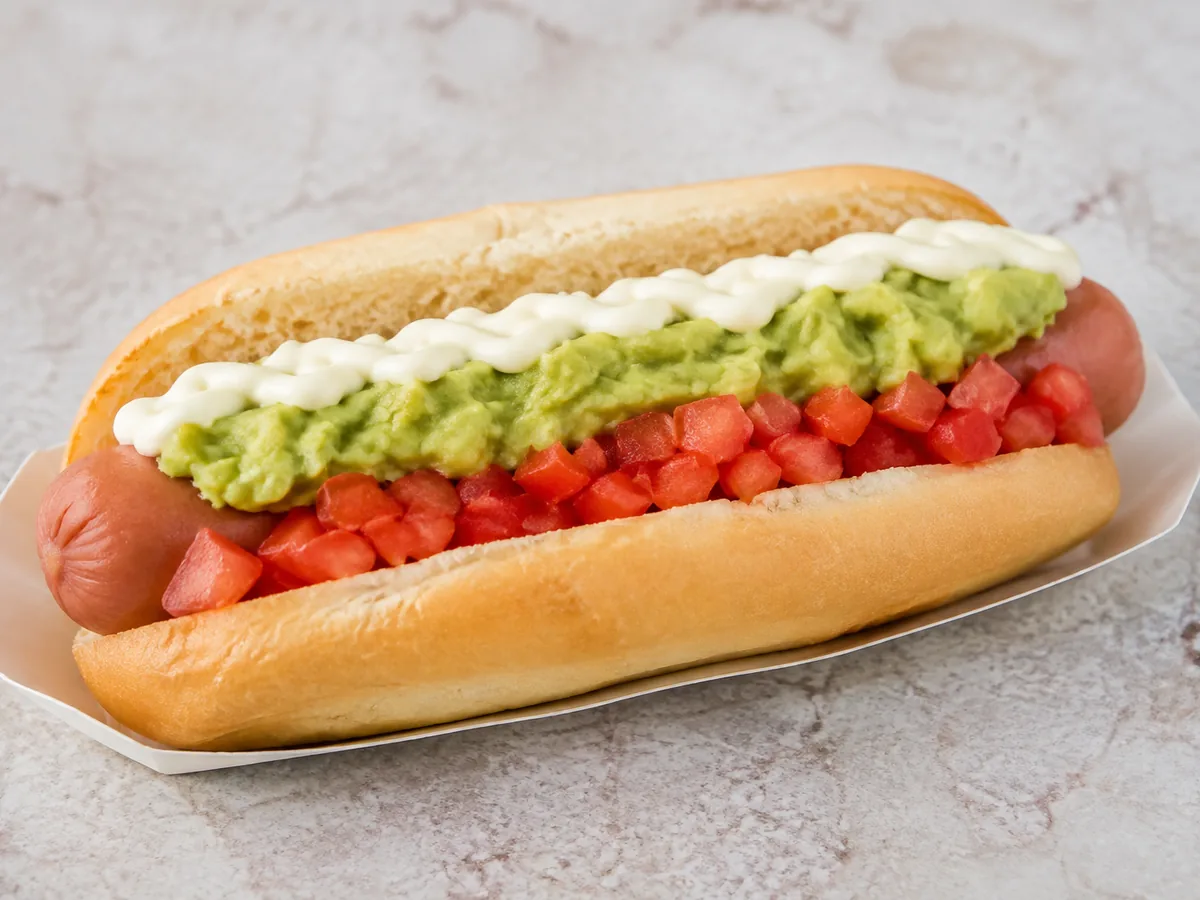 A long soft bun on a white paper tray holding a pink sausage under a band of finely diced red tomato, a band of mashed green avocado and a piped white crest of mayonnaise along its length.
