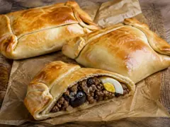 Three large rectangular golden baked empanadas with thick folded seams on parchment, the front one cut open to show diced beef, onion, a black olive and a wedge of hard-boiled egg.