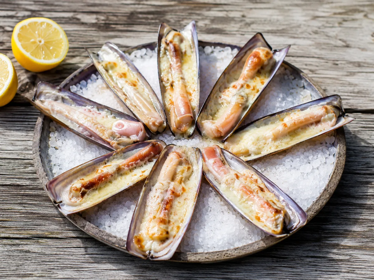 A round platter of coarse white salt holding purple-edged razor clam shells arranged in a wheel, each filled with a pale clam under browned melted cheese, with two lemon halves on the weathered wood beside it.