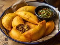 A blue-rimmed plate of deep-yellow fried half-moon empanadas on parchment, one broken open to show a filling of browned mince and diced potato, with a small black bowl of chopped green and red ají beside them.