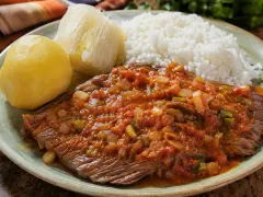 A wide slab of braised beef under a chunky red sauce of tomato, onion and green herbs, on a green-rimmed plate with white rice, a piece of boiled yuca and a round yellow potato alongside.