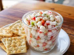 A clear glass cup packed with small snow-white cubes of cured fish flecked with red pepper, green celery and cilantro in cloudy citrus juice, with square soda crackers fanned on the plate beside it.
