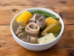 A deep white bowl of clear golden broth holding a round marrow bone, chunks of beef, a wheel of yellow corn on the cob, green beans, chayote, sweet potato and cassava, on a rustic wooden table.