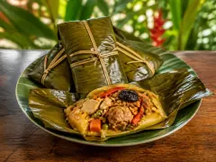 Two green banana-leaf parcels tied with string standing behind an opened tamal on a leaf-lined plate, showing yellow masa filled with pork, rice, carrot, peas, a red pepper strip and a dark prune.