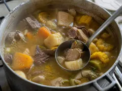 A deep bowl of Cuban ajiaco — chunks of yuca, calabaza, corn on the cob and meat in a thick gold-orange broth.