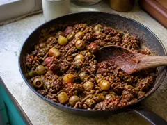 A skillet of Cuban picadillo — browned ground beef in rust-red tomato sauce with green olives and plump raisins — beside white rice.