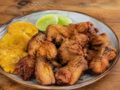 A blue-grey rimmed plate heaped with craggy golden-brown fried bone-in chicken pieces, with two halved limes behind them and three flat golden tostones fanned along the left edge, on a wooden table.