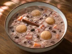 A wide bowl of pale rosy-pink creamy dessert holding whole dark red beans, orange sweet potato cubes and raisins, with a cinnamon quill laid across it and four round pale milk cookies floating on the surface.