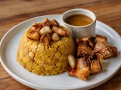 A pale ceramic plate holding a golden dome of coarsely mashed plantain studded with browned pork pieces and whole garlic cloves, beside a small cup of amber broth and a heap of crisp chunks of fried pork belly.