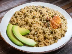 A speckled white bowl heaped with pale golden rice studded with olive-green pigeon peas, a piece of golden-brown toasted rice crust on top, and three slices of avocado fanned along one side, on a wooden table.