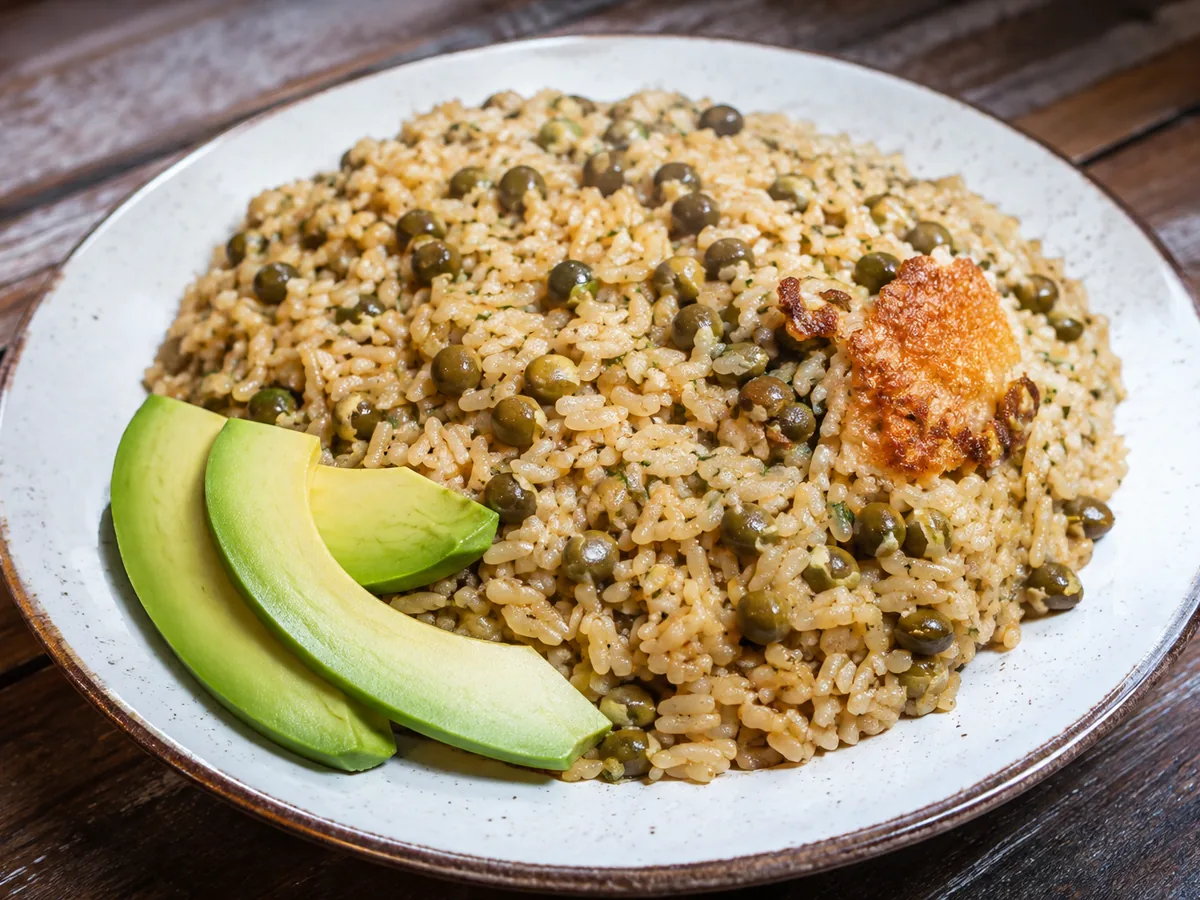 A speckled white bowl heaped with pale golden rice studded with olive-green pigeon peas, a piece of golden-brown toasted rice crust on top, and three slices of avocado fanned along one side, on a wooden table.