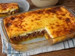 A glass baking dish of layered casserole with a blistered golden-brown cheese top, a corner cut away to show a pale plantain top layer, a thick band of dark minced beef and a yellow plantain base, on a blue-checked cloth.