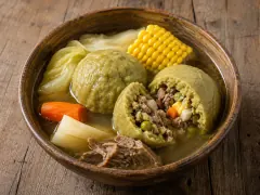 A wide bowl of clear amber beef broth holding a large smooth golden plantain dumpling cut open to show its beef and egg filling, with a round of corn on the cob, yuca chunks and shredded cabbage alongside.