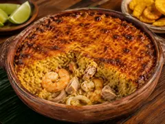 A round dark clay dish of golden-brown baked casserole with a toasted blistered surface, a portion scooped away to reveal pink shrimp and white fish beneath, with lime wedges and plantain chips alongside.