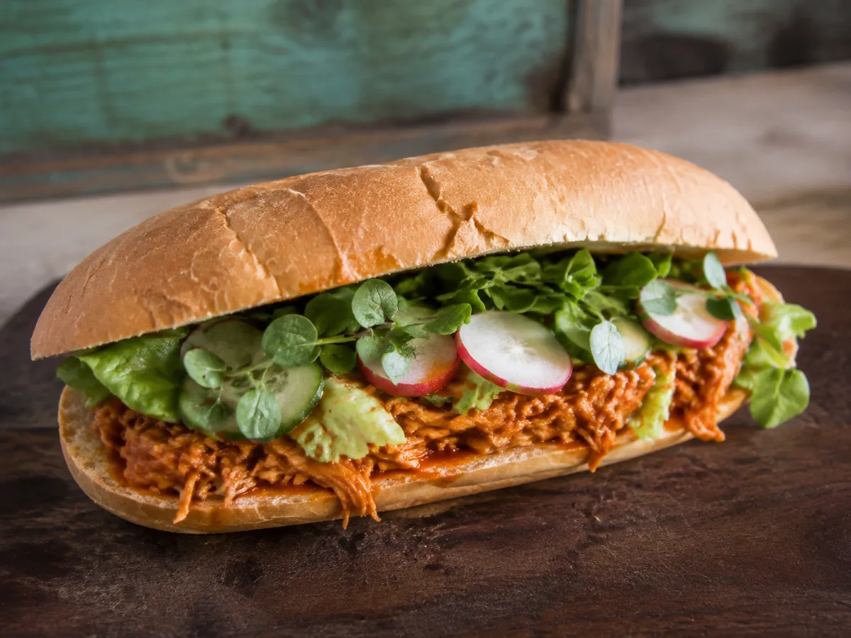 A long crusty roll packed with shredded chicken in orange-red sauce, sliced cucumber, radish coins and green watercress, on a dark wooden board.