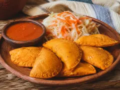 Five orange fried corn half-moons with crimped edges on a wooden plate, with a bowl of red sauce and a heap of shredded cabbage and carrot slaw behind.