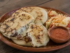 Three pale corn cakes with dark toasted blisters on a terracotta plate, one torn open to show melting white cheese and dark filling, with shredded cabbage slaw and a bowl of red salsa alongside.