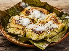 Four pale golden corn cakes with dark griddle marks on a charred green banana leaf in a terracotta dish, topped with white cream and crumbled cheese.