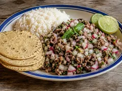 A blue-rimmed platter of finely minced beef flecked with white onion, pink radish and green herbs, topped with a mint sprig, beside white rice, lime wedges and stacked corn tortillas.