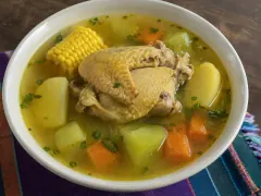 A white bowl of clear golden broth holding a bone-in chicken piece with yellow skin, corn on the cob, potato, chayote and carrot chunks, on a striped woven cloth.