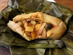 An opened banana leaf holding a pale golden tamal split to show shredded chicken, potato pieces, a green olive and red sauce inside.