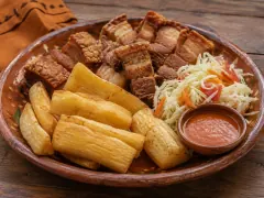 A terracotta platter of golden fried cassava batons with thick pieces of fried pork belly, a heap of cabbage and carrot slaw and a small bowl of red sauce.