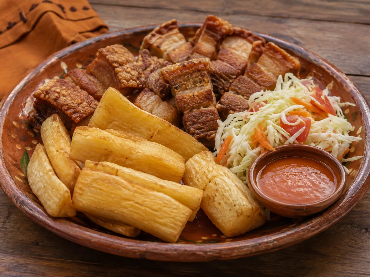 A terracotta platter of golden fried cassava batons with thick pieces of fried pork belly, a heap of cabbage and carrot slaw and a small bowl of red sauce.
