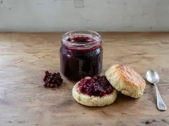 An open jar of dark red berry jam, a split scone spread with the jam, a sprig of small red berries and a teaspoon on a wooden table.